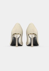 Beige high-heeled shoes with a pointed toe, smooth leather upper, and metallic gold accents on the heel. Black sole with branding detail.