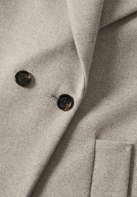 Gros plan sur un manteau en laine gris clair montrant deux boutons foncés et une poche sur un tissu texturé.