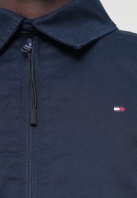 Marineblå zip-up jakke med krave, prydet med en sort lynlås og et lille broderet logo i rød, hvid og blå på venstre bryst.