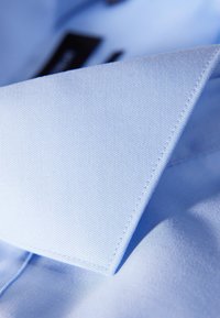 Colletto di camicia azzurro chiaro con una texture liscia, angoli definiti e un dettaglio di cucitura ordinato. Include etichette visibili vicino al colletto.