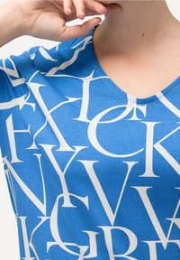 Blaues T-Shirt mit kurzen Ärmeln und einem weißen abstrakten Buchstabenprint. V-Ausschnitt und weiches Baumwollmaterial. Nahaufnahme der Stoffstruktur und des Designs.