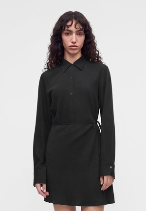 Femme aux cheveux frisés et foncés portant une robe noire à manches longues avec col et boutons, debout devant un fond clair uni.