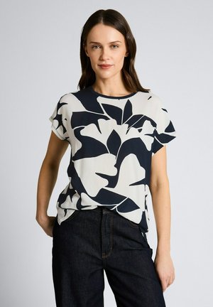 Młoda kobieta w krótkim rękawie z czarno-białym kwiatowym wzorem oraz ciemnych dżinsach, stojąca na tle jednolitego jasnego tła.