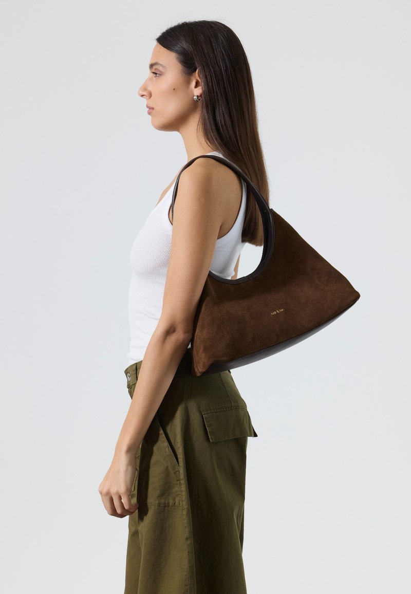 Jeune femme en débardeur blanc et pantalon olive portant un sac bandoulière en daim marron avec des finitions noires, debout de profil sur un fond uni.