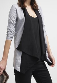 Grauer langärmeliger Cardigan mit einem drapierten schwarzen Vorderteil. Weicher Stoff, offenes Design, kombiniert mit einem schwarzen Oberteil und schwarzen Hosen. Leicht strukturiert.