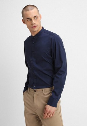 Marineblaue Langarm-Hemdbluse mit Stehkragen, glattem Stoff, Frontknöpfen und tailliertem Schnitt, kombiniert mit beigen Hosen.