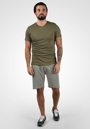 Maslinasto zelena majica kratkih rukava s suptilnom teksturom, uparena sa svijetlosivim kratkim hlačama. Outfit je upotpunjen tamnoplavim sportskim cipelama.