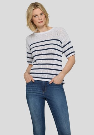 Frau mit kurzem blondem Haar, die ein weißes Strickoberteil mit marineblauen Streifen und blaue Jeans trägt, steht mit den Händen in den Taschen vor grauem Hintergrund.