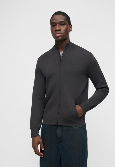 Άνδρας που φοράει σκούρο γκρι ζακέτα με φερμουάρ και τσέπες και μπλε τζιν, στέκεται μπροστά σε απλό ανοιχτό γκρι φόντο.