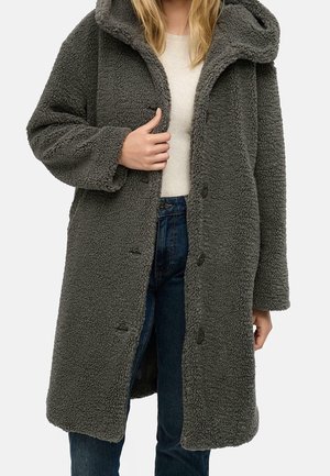 Femme portant un long manteau polaire texturé gris foncé sur un pull blanc et un jean bleu, tenant le manteau ouvert d'une main.