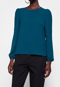 Donna che indossa una blusa lunga a maniche in teal con spalle a sbuffo e polsini elastici, abbinata a pantaloni cargo neri su sfondo bianco.