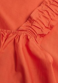 Oranje stof met een gladde textuur, met geplooide accenten en gestikte details langs de naden. Close-up van het materiaal en het ontwerp.