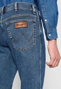 Close-up van een persoon die blauwe denim Wrangler jeans en een bijpassend denim overhemd met drukknoopmanchet draagt, waarbij de achterzak en rechterhand te zien zijn.
