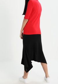 Žena v červeném topu, asymetrické černé sukni a bílých teniskách, stojící s jednou nohou mírně vpřed na bílém pozadí.