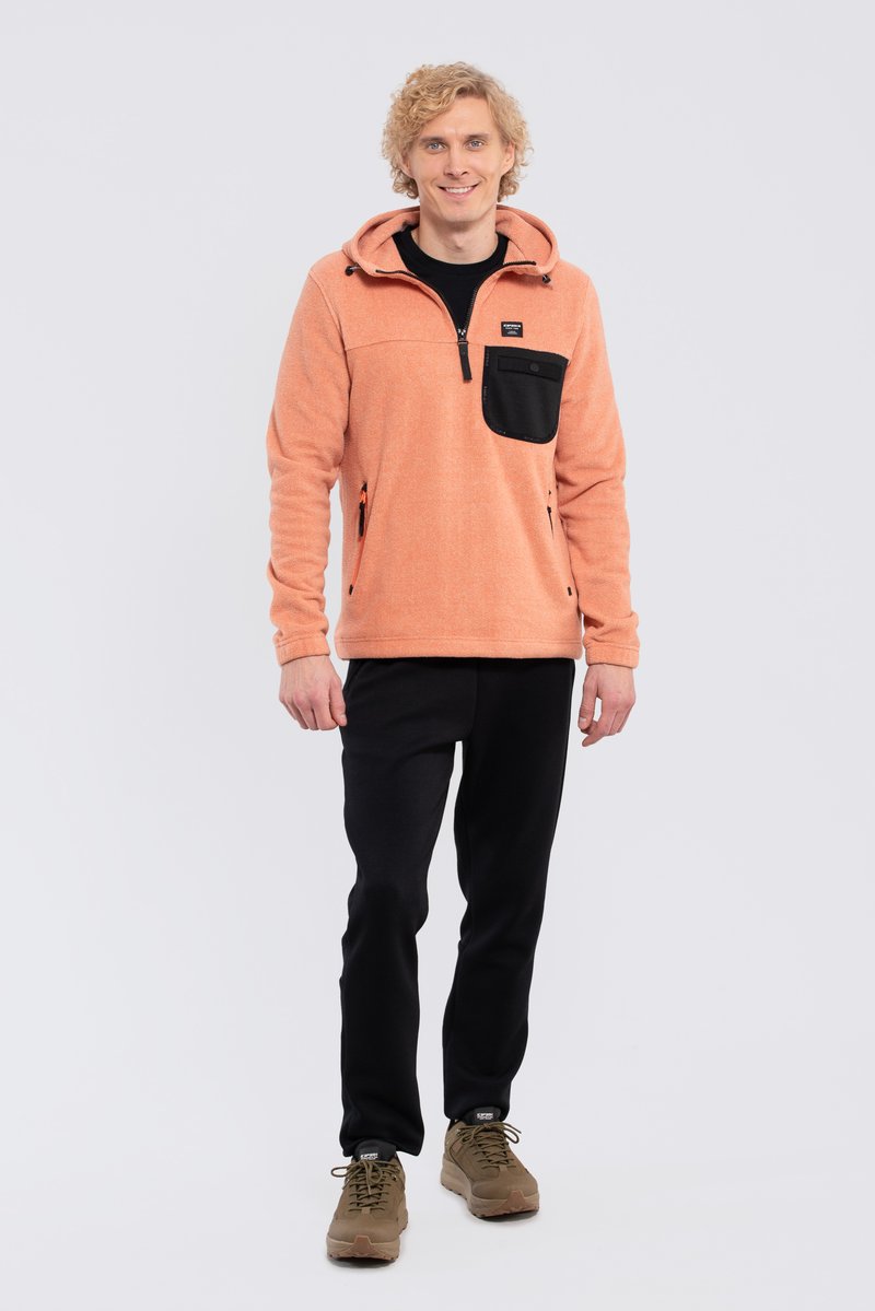 Fleece pullover i koralfarve med en sort lynlås lomme, to sidelommer med lynlås og en justerbar hætte. Sorte bukser og beige sneakers.