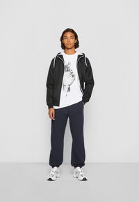 Čierny vetrovka s bielymi akcentmi, grafické biele tričko s otlačkom ruky, námornícke tepláky a biele tenisky. Prezentácia ležérneho outfitu.