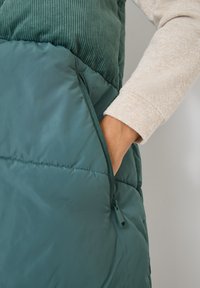 Türkisfarbener, plüschiger Weste aus glattem Stoff mit einem Oberteil aus Baumwollcord. Verfügt über eine seitliche Reißverschlusstasche und einen weichen, strukturierten Ärmel.