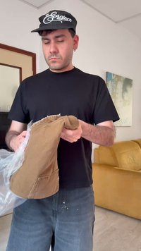 Άνδρας που φοράει μαύρη μπλούζα και καπέλο ξετυλίγει ένα ζευγάρι διπλωμένα καφέ παντελόνια σε εσωτερικό χώρο κοντά σε ένα κίτρινο καναπέ και αφηρημένη τοιχογραφία.