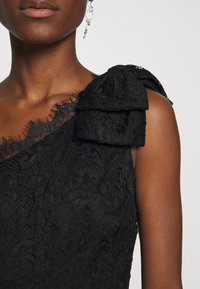 Robe en dentelle noire avec une conception asymétrique à une épaule, un détail à volants sur l'épaule et des motifs floraux complexes sur tout le tissu.