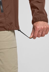 Brown lichte jas met een trekkoord aan de onderrand, voorzien van een ritszak en elastische manchetten. Trek aan het trekkoord voor aanpassing.