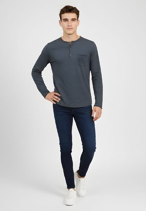 Joven de pie con la mano en la cadera, vestido con camisa henley de manga larga oscura, jeans ajustados oscuros y zapatillas blancas sobre fondo blanco.