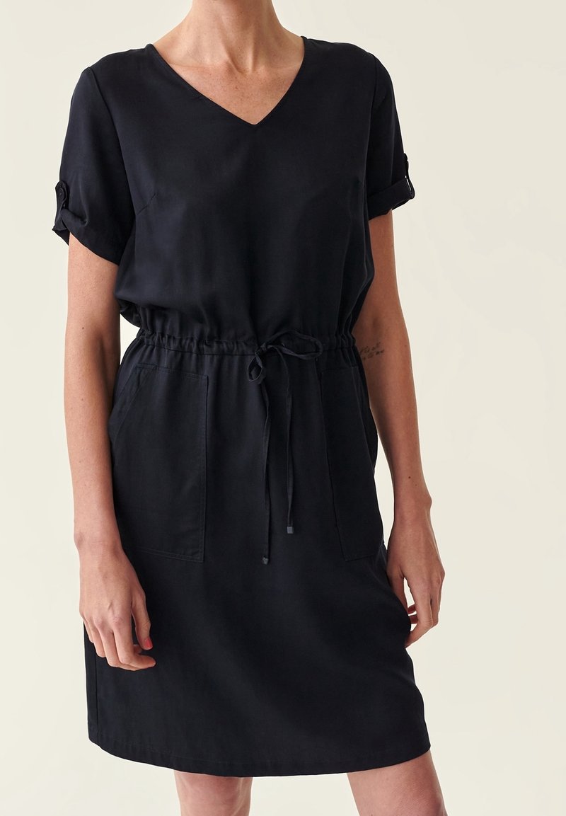 Femme portant une robe noire à manches courtes avec col en V, taille à cordon, manches retroussées et deux poches avant, sur un fond clair uni.