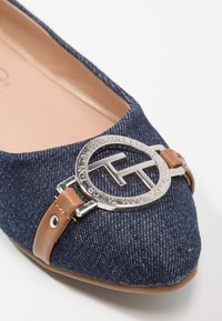Gros plan d'une chaussure plate en denim bleu marine avec une bride en cuir marron et une boucle argentée circulaire avec le logo Tom Tailor sur le bout.