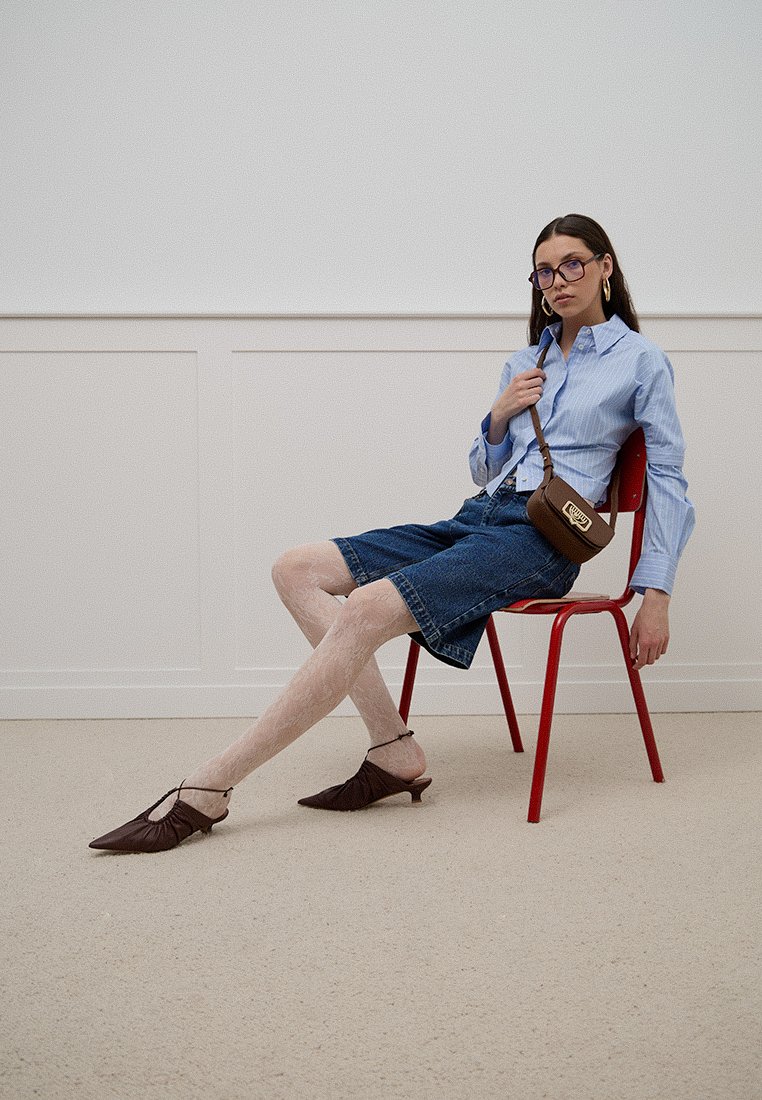 Zapatos planos de cuero marrón, camisa de botones a rayas azules, pantalones cortos de mezclilla y un bolso bandolera marrón. Colocados sobre una silla roja contra una pared blanca.