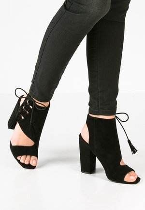 Benen in zwarte skinny jeans met zwarte suède enkellaarsjes met open teen, blokhak, veters en franjes.