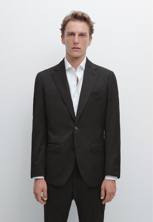 Mand med kort blondt hår iført en mørkegrå jakkesætjakke over en hvid skjorte, stående foran en ensfarvet lys baggrund.