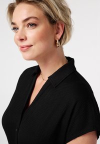 Chemise noire à col, en tissu texturé, avec des manches courtes et un détail de bouton. Des boucles d'oreilles en or et un collier délicat en chaîne sont portés.
