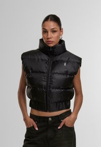 Black padded vest with a high collar and sleeveless design, featuring a front zipper and textured material. A logo is visible on the chest.