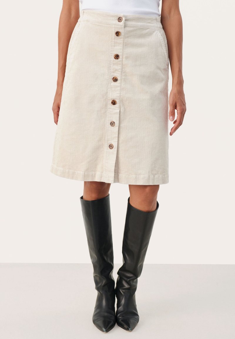 Kvinde iført en beige kordnøgle nederdel med knapper foran kombineret med sorte knælange læderstøvler mod en ensfarvet baggrund.