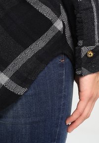 Mano appoggiata accanto a jeans blu, indossando una camicia a fantasia nera e grigia con tessuto testurizzato e un pulsante di legno sul polsino.