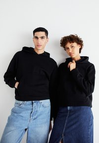 Sorte hettegensere med frontlommer, båret av to modeller. Den ene har på seg lyse jeans, mens den andre har en denimskjørt med sidedeling.