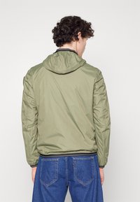 Chaqueta ligera de color verde oliva con capucha, puños elásticos y dobladillo, con una textura suave y un diseño minimalista. Lleva puesta sobre unos vaqueros azules.