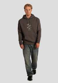 Dunkelgrauer Hoodie mit einer Frontillustration eines Charakters, kombiniert mit hellblauen Jeans und schwarzen Turnschuhen. Lässiger, bequemer Schnitt.