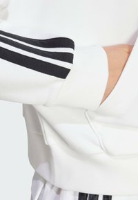 Weißes Sweatshirt mit schwarzem Drei-Streifen-Design auf dem Ärmel, mit geripptem Look und Kängurutasche auf der Vorderseite.