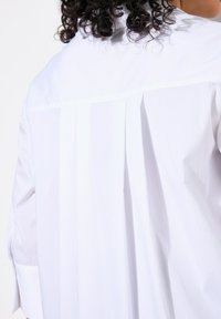 Camicia bianca con un design strutturato, caratterizzata da una piega sul retro per una maggiore texture e movimento. Tessuto di cotone liscio, maniche lunghe.