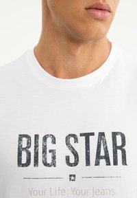 Primo piano di una persona che indossa una maglietta bianca con grandi scritte nere usurate "BIG STAR" e testo più piccolo "Your Life. Your Jeans."