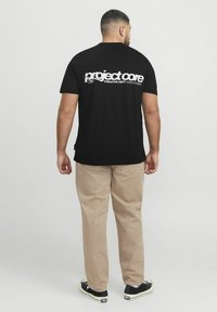 Camiseta de algodón negra con texto blanco en la parte trasera: "departamento creativo de PROJECT CORE." Combinada con pantalones beige y zapatillas negras.