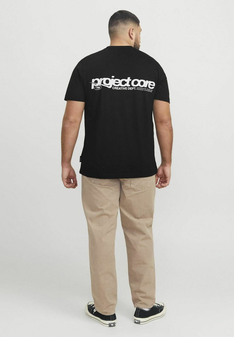 Camiseta de algodón negra con texto blanco en la parte trasera: "departamento creativo de PROJECT CORE." Combinada con pantalones beige y zapatillas negras.