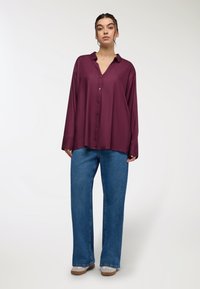Camicia bordeaux con colletto, maniche lunghe e vestibilità comoda, abbinata a jeans a gamba larga azzurri e scarpe beige.