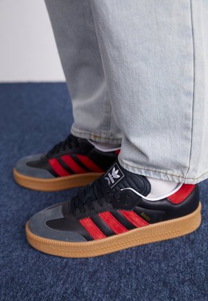 Personne portant des baskets Adidas Samba noires et rouges avec des semelles en gomme et un jean bleu clair, debout sur un tapis bleu.