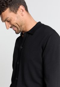 Zwarte cardigan van een gestructureerde brei stof, met een knoopsluiting aan de voorkant, kraag en een slanke pasvorm.