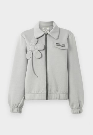 Lys grå zip-jakke med blomstermotiv på venstre side, elastiske mansjetter og krage, med et merkelapp på brystlommen.