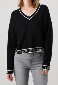 Pull court noir avec col en V bordé de blanc, manches longues, et logo de la marque tissé sur l'ourlet et les poignets. Associé à un jean gris.