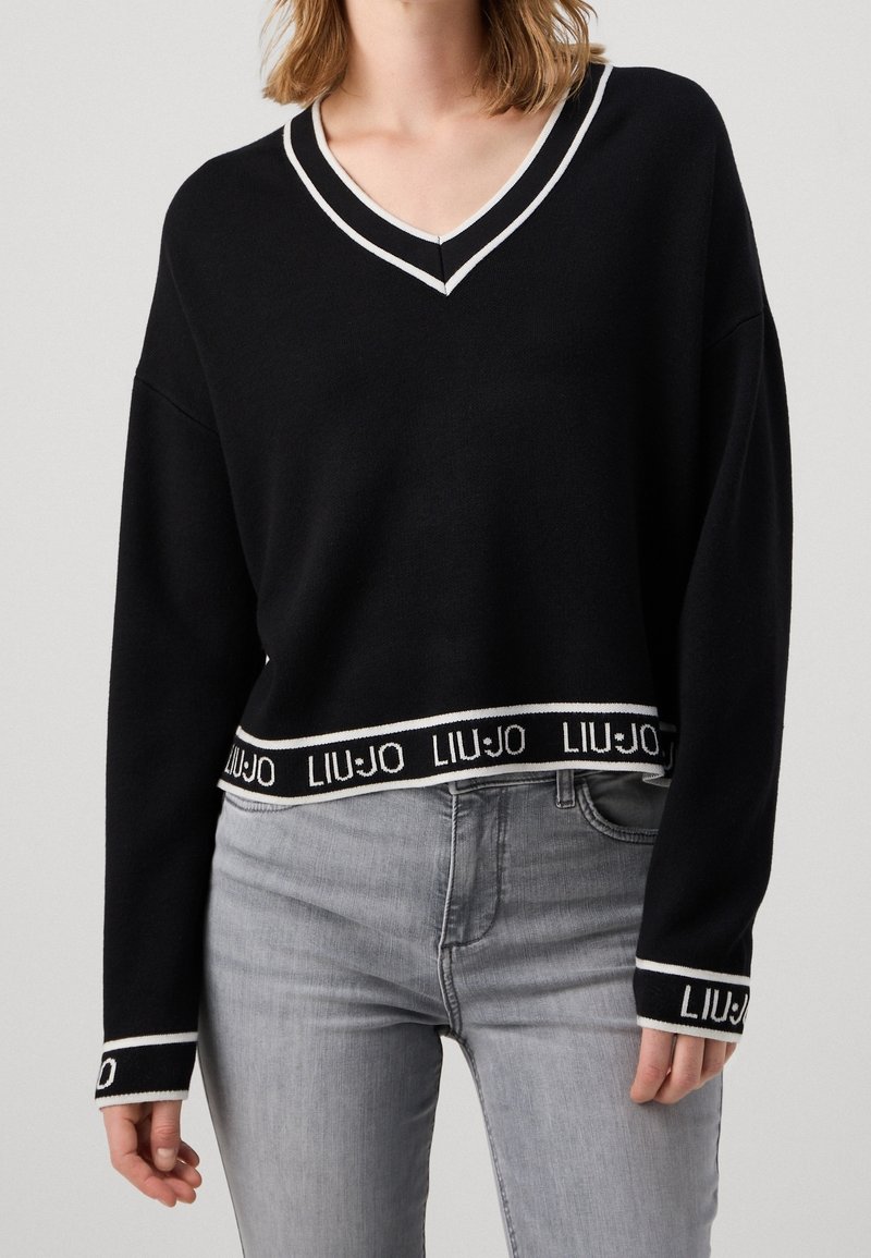 Pull court noir avec col en V bordé de blanc, manches longues, et logo de la marque tissé sur l'ourlet et les poignets. Associé à un jean gris.