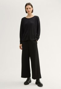 Schwarzes Langarmoberteil mit lockerer Passform, ergänzt durch schwarze weite Hosen und klobige schwarze Ankle Boots. Glatte Stofftextur.