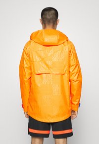 Ljusstark orange vattentät jacka med huva. Har ett upprepat logomönster, långa ärmar och en avslappnad passform. Kombinerad med svarta kortbyxor.